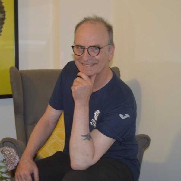 A picture of a man sat down on a chair, wearing glasses, posing and smiling.