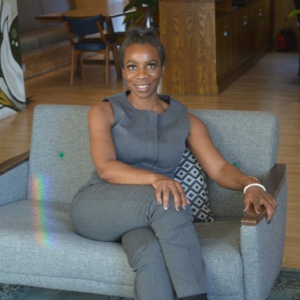 Black British female sat down on a grey sofa, smiling, crossed legs, wearing a grey suit.