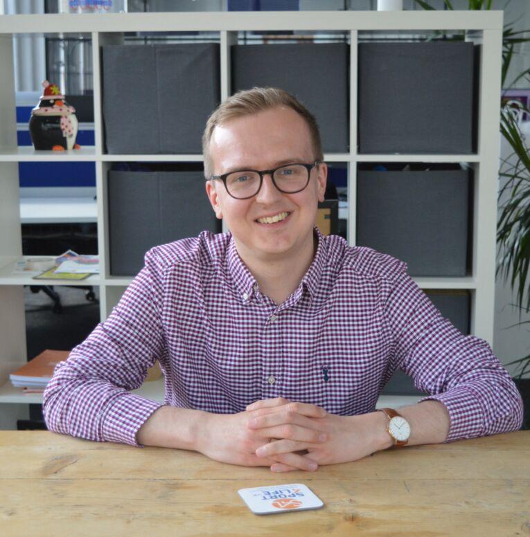 White male, sat down, resting arms on a wooden table, wearing glasses and a checked shirt.