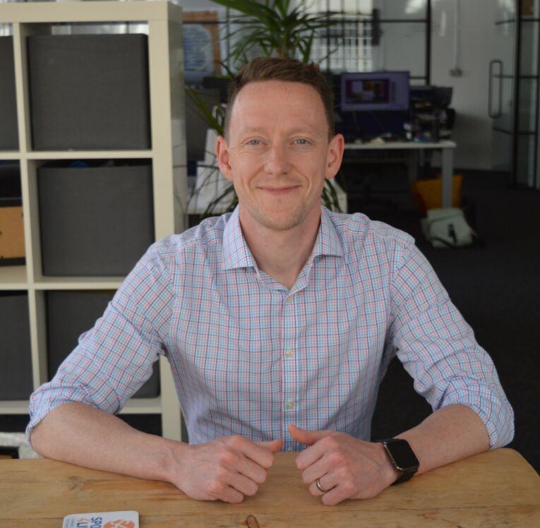 White male, sat down, smiling, resting arms on a wooden table, wearing a checked light shirt.