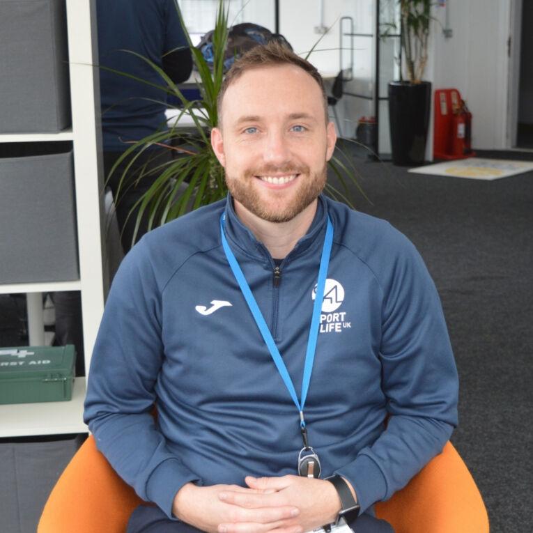 White male with facial hair, smiling, sat on an orange chair, wearing Sport 4 Life uniform.